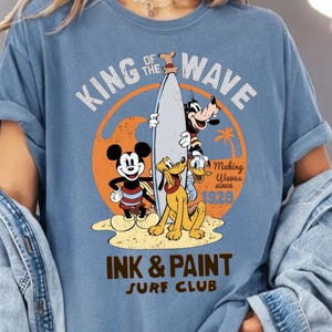 May include: Blue t-shirt featuring Mickey Mouse, Goofy, and Pluto with a surfboard. The shirt has the text "King of the Wave" and "Ink & Paint Surf Club". The design includes an orange wave and palm tree.