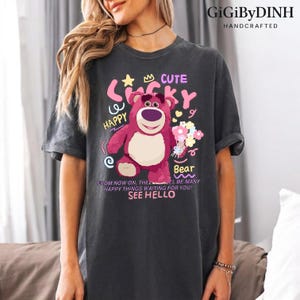 A woman wearing a gray t-shirt with a colorful graphic of a pink bear and various text elements, including the words 'Cute', 'Happy', 'Bear', and 'See Hello'.