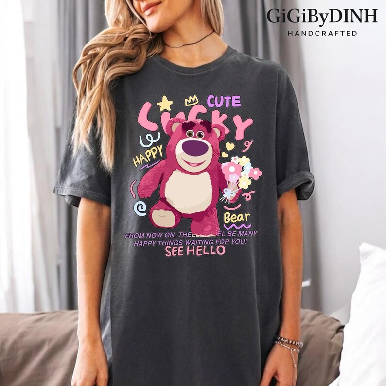 A woman wearing a gray t-shirt with a colorful graphic of a pink bear and various text elements, including the words 'Cute', 'Happy', 'Bear', and 'See Hello'.