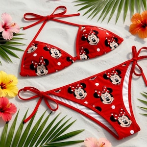 May include: Red bikini set featuring a Minnie Mouse pattern with white polka dots. The top and bottom are tied with red strings. The image includes the text "GiGiByDinh Handcrafted" and is surrounded by tropical flowers and palm leaves.