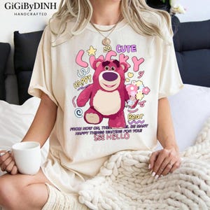 A woman wearing a white t-shirt with a cartoon image of a pink bear and text that says "Cute Happy Bear" and "From now on, the waiting for you is many happy things see hello".