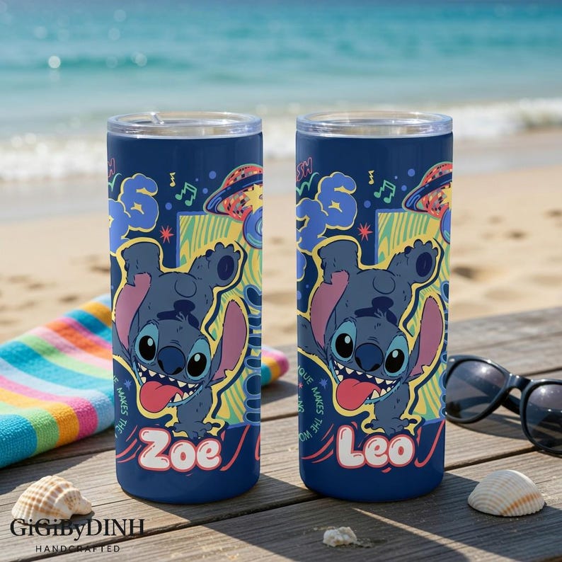 Two personalized tumblers with a cartoon character design, placed on a wooden surface near a beach towel and sunglasses.