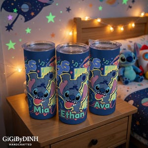 Three personalized tumblers with the character Stitch's face printed on them, each with a different name: "Mia", "Ethan", and "Ava". The tumblers are placed on a wooden dresser, with a stuffed animal and a string of lights visible in the background.