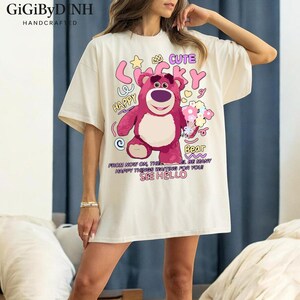 A woman wearing a white t-shirt with a cartoon image of a pink bear and various text and graphics printed on it.