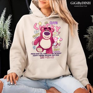 A person wearing a white hoodie with a cartoon image of a pink bear and text that says "Cute Lucky Happy Bea" printed on it.