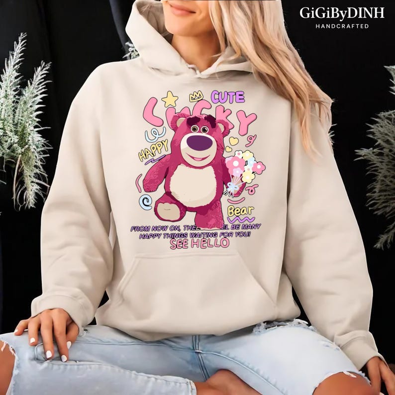 A person wearing a white hoodie with a cartoon image of a pink bear and text that says "Cute Lucky Happy Bea" printed on it.