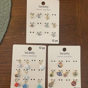 May include: Three sets of "bead landing" charms on white cards. The charms include dinosaurs, dice, and eye designs. Each card is labeled "12 pc".