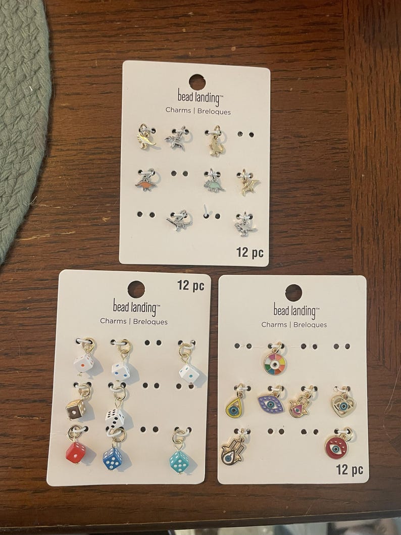 May include: Three sets of "bead landing" charms on white cards. The charms include dinosaurs, dice, and eye designs. Each card is labeled "12 pc".