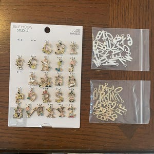 May include: A collection of gold-toned and white letter charms with floral accents. The charms are displayed on a card from Blue Moon Studio, along with two small bags of additional letters. The image shows a variety of decorative alphabet charms.
