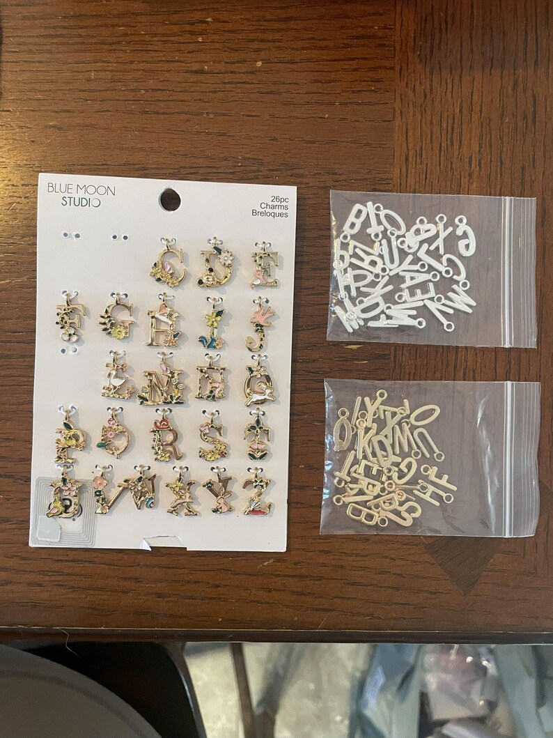 May include: A collection of gold-toned and white letter charms with floral accents. The charms are displayed on a card from Blue Moon Studio, along with two small bags of additional letters. The image shows a variety of decorative alphabet charms.
