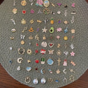 May include: A collection of small, decorative charms in various shapes and colors, including hearts, flowers, musical notes, and stars. The charms are made of metal and some have colorful enamel accents. They are arranged on a green, textured surface.