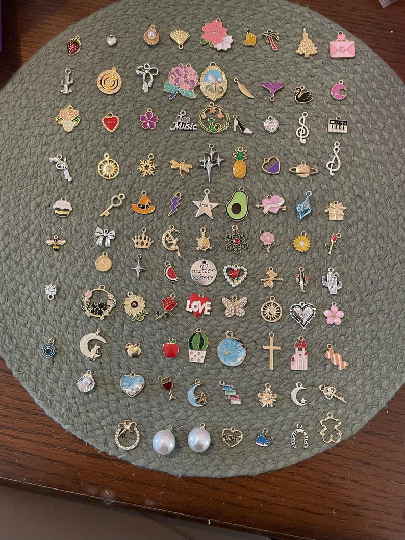 May include: A collection of small, decorative charms in various shapes and colors, including hearts, flowers, musical notes, and stars. The charms are made of metal and some have colorful enamel accents. They are arranged on a green, textured surface.