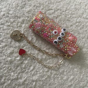 May include: A bedazzled pink and gold lipstick case with the word "MORE" spelled out in white and black beads. A gold chain with a heart-shaped lock and a red heart charm is attached. The case is covered in rhinestones and decorative elements.