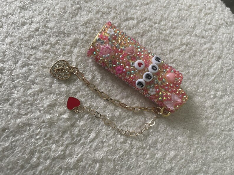 May include: A bedazzled pink and gold lipstick case with the word "MORE" spelled out in white and black beads. A gold chain with a heart-shaped lock and a red heart charm is attached. The case is covered in rhinestones and decorative elements.