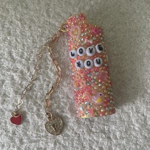 May include: A bedazzled pink lighter case with the words "LOVE YOU" spelled out in white letter beads. The case is covered in pink and clear rhinestones, with a gold chain and heart charms attached.