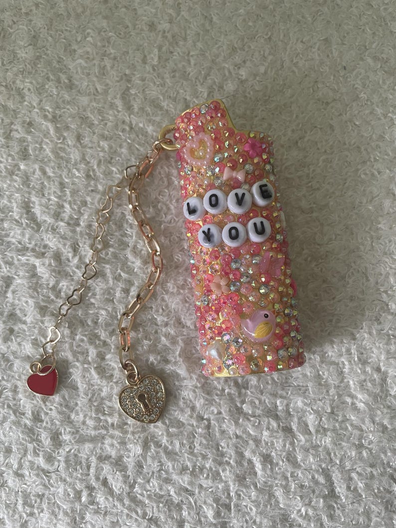 May include: A bedazzled pink lighter case with the words "LOVE YOU" spelled out in white letter beads. The case is covered in pink and clear rhinestones, with a gold chain and heart charms attached.