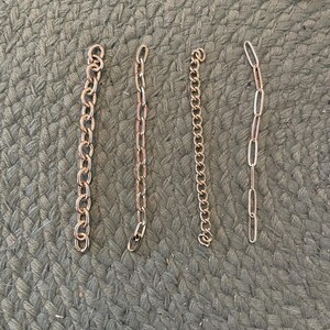 May include: Four gold-colored metal chains of varying styles and lengths are arranged on a textured, green surface. The chains include a curb chain, a cable chain, a rolo chain, and an oval link chain. A small, round, floral-themed pendant is in the upper right corner.