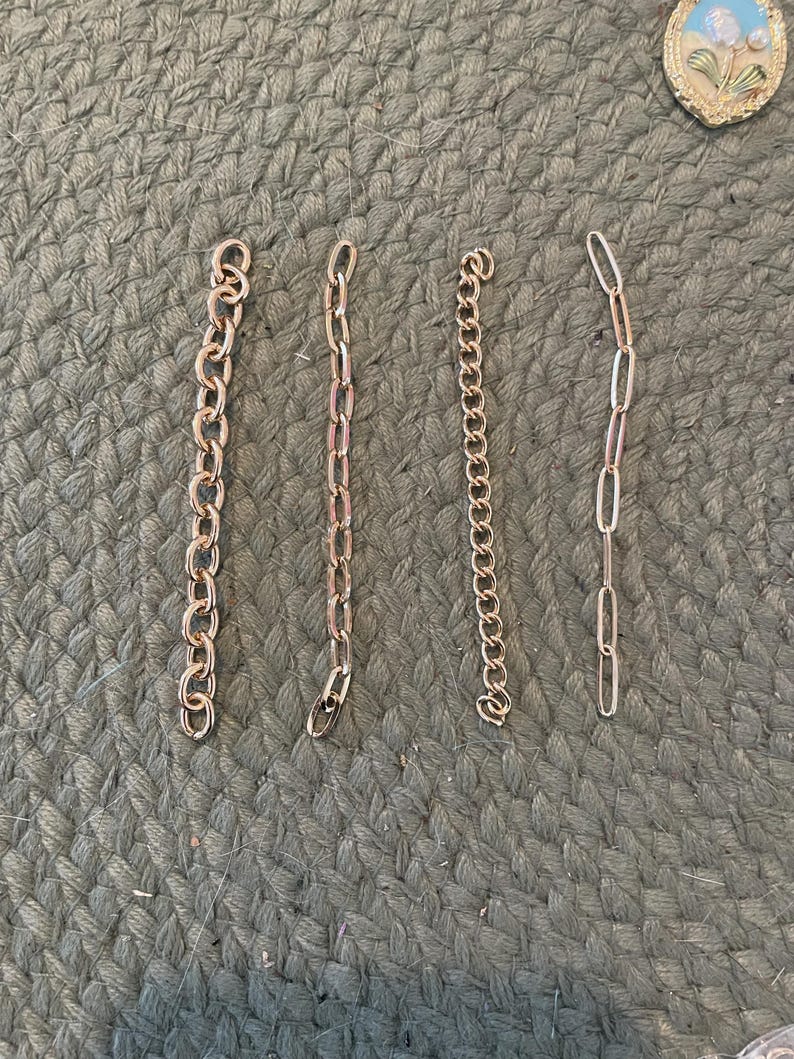 May include: Four gold-colored metal chains of varying styles and lengths are arranged on a textured, green surface. The chains include a curb chain, a cable chain, a rolo chain, and an oval link chain. A small, round, floral-themed pendant is in the upper right corner.