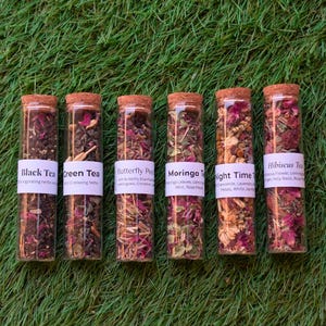 May include: Six glass tubes filled with different herbal teas, each sealed with a cork stopper. The teas include Black Tea, Green Tea, Butterfly Pea, Moringa Tea, Night Time, and Hibiscus Tea. Each tube has a label with the tea's name and ingredients.