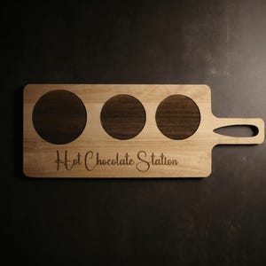 Könnte beinhalten: Holzservierbrett mit drei kreisförmigen Aussparungen und einem Griff. Auf dem Brett steht in kursiver Schrift der Schriftzug "Hot Chocolate Station". Die Aussparungen sind dunkelbraun, das Brett hellbraun.