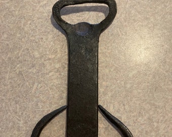 May include: A black metal bottle opener with a unique design. The opener has a traditional bottle opener at the top and two hooks at the bottom.