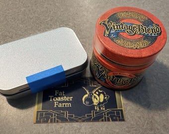 May include: A rectangular silver metal tin with a blue tape strip, a cylindrical red metal container labeled Vintage Blend, and a rectangular dark blue card featuring a yellow sheep illustration and the text Fat Toaster Farm.
