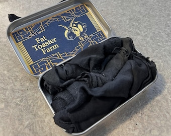 May include: A rectangular silver metal tin containing folded black fabric. The inside of the lid features a blue and gold graphic with the text Fat Toaster Farm and an illustration of a sheep with large horns, framed by geometric gold lines.