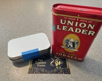 May include: A rectangular silver metal tin with a blue tape strip, a red Union Leader smoking tobacco tin with an eagle graphic, and a rectangular card featuring the text Fat Toaster Farm and a stylized goat illustration in gold and black.