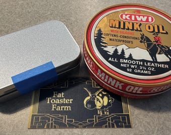 May include: A rectangular silver metal tin with a blue tape strip, a round Kiwi Mink Oil tin with a red and gold lid featuring a horse and rider graphic, and a rectangular card with the text Fat Toaster Farm and a stylized sheep illustration.