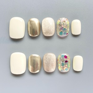 May include: A set of ten press-on nails arranged in two rows. The nails feature a variety of finishes including solid cream, metallic gold, fine champagne glitter, and clear acrylic with multi-colored chunky glitter accents.