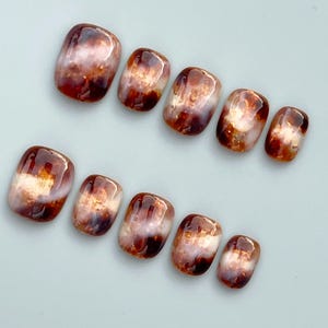 May include: A set of ten press-on nails featuring a glossy, marbled finish in shades of deep mahogany, amber, and translucent white. Each nail has a rounded rectangular shape with a smooth, reflective surface and blended colour patterns.