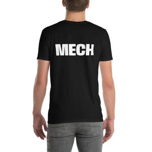 Op de afbeelding: Zwart T-shirt met het woord "MECH" in grote, witte hoofdletters op de rug gedrukt. Het shirt heeft korte mouwen en lijkt van een zacht materiaal te zijn. De persoon draagt een grijze jeans.