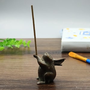 May include: A bronze rabbit incense holder, holding a brown incense stick. The rabbit is depicted with its arms raised and ears perked. The holder is on a wooden surface. The incense stick is approximately 20 cm long.