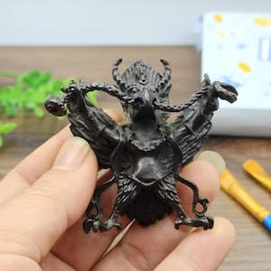 Può includere: Statuetta in bronzo scuro di una creatura mitica con testa di uccello, corna e ali spiegate. La figura presenta dettagli intricati, tra cui un volto dettagliato, piume e piedi artigliati. La statuetta è tenuta in una mano.