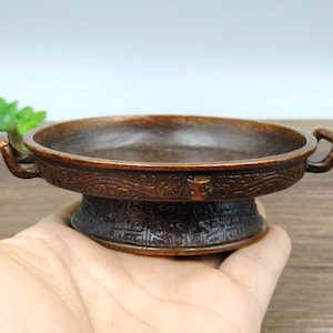 Pode incluir: Uma tigela de cor bronze, de estilo antigo, com duas alças. A tigela tem uma bacia larga e rasa e uma base decorativa com padrões. A tigela é segurada na mão de uma pessoa.
