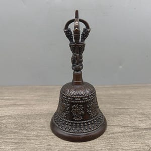 Puede incluir: Una campana de color bronce con intrincadas tallas y un asa decorativa. La campana presenta patrones detallados y un asa con un diseño curvo. La campana es probablemente de metal y tiene una apariencia vintage o antigua.