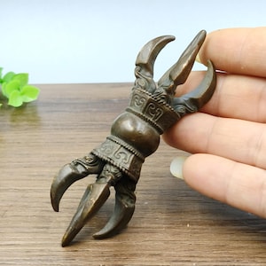 May include: A bronze-coloured claw-shaped sculpture with detailed patterns. The sculpture has a central bulbous section with ornate designs and three curved, pointed claws. The piece is held in a hand, indicating its size and scale.