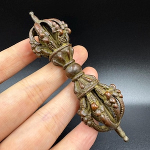 May include: A bronze-coloured vajra, a ritual object, is held in a hand against a dark background. The vajra features intricate details, including a central sphere, ornate floral designs, and a crown-like top. The object is approximately 15 cm long.