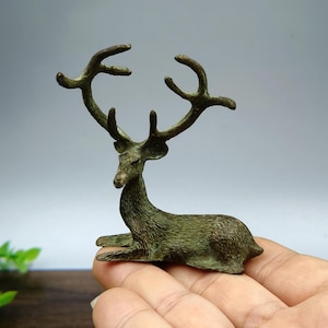 May include: A bronze-coloured deer figurine in a resting pose. The deer has large antlers and is lying down with its legs tucked beneath its body. The figurine is held in a hand, showcasing its size and detail. The deer has a textured surface and a detailed face.