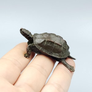 May include: A small, dark bronze-coloured turtle figurine with a detailed, textured shell and visible scales on its legs and head. The sculpture is cast in a realistic pose with the head extended forward and the limbs positioned as if walking.