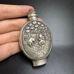 May include: Antique silver-colored snuff bottle with a detailed relief design. The bottle features a scene with two figures and a bear, surrounded by trees and flowers. The stopper is red.