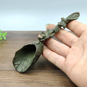 May include: A bronze-colored decorative spoon with a leaf-shaped bowl and a handle adorned with floral and botanical details. The spoon is held in a hand, showcasing its intricate design and craftsmanship. The spoon's overall aesthetic suggests a vintage or antique style.
