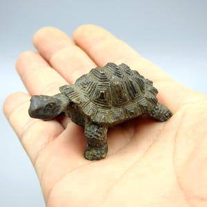 May include: A small, bronze-colored turtle figurine with a detailed shell pattern. The turtle is held in an open hand, showcasing its size and intricate design. The figurine has a weathered appearance, suggesting an antique or vintage aesthetic.