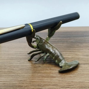 Könnte beinhalten: Ein schwarzer Stift mit goldenen Akzenten liegt auf einem bronzefarbenen, hummerförmigen Stifthalter. Der Stifthalter ist detailliert mit den Scheren und dem Körper des Hummers gestaltet, was ihn zu einem einzigartigen Schreibtischaccessoire macht.