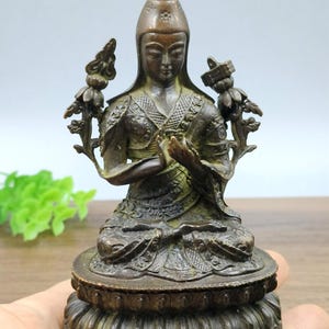 May include: A bronze statue of a seated figure in a meditative pose. The figure wears a pointed hat and ornate robes, with hands clasped in prayer. Decorative floral elements flank the figure. The statue is mounted on a detailed lotus base.