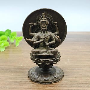Puede incluir: Estatua de color bronce de una deidad de múltiples brazos sentada sobre una base de flor de loto, con un halo circular detrás de la cabeza. La estatua tiene detalles intrincados y se exhibe sobre una superficie de madera. La estatua es una representación de una figura religiosa.