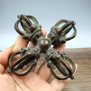 Könnte beinhalten: Ein bronzefarbener Vajra, ein rituelles Objekt, das in einer Hand gehalten wird. Das symmetrische Design weist vier gebogene Zacken auf, die sich von einer zentralen Kugel erstrecken, mit aufwendigen Details und einem verwitterten Aussehen. Das Objekt wird wahrscheinlich in buddhistischen Praktiken verwendet.