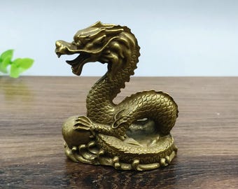 May include: A detailed brass dragon figurine with an open mouth and a textured body. The dragon is coiled around a sphere, showcasing intricate craftsmanship. The figurine is a golden-brown color.