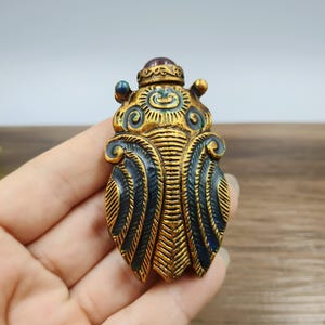 May include: A detailed, gold-colored decorative beetle with blue and green accents. The beetle has a textured surface and a small, red gem on top. The wings are detailed with a feather-like pattern.