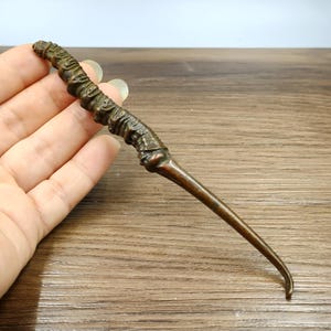 May include: A bronze-colored wand with a textured handle and a slender, curved tip. The wand's handle has a detailed, organic design. The wand is held in a hand, suggesting its use as a magical item or prop.